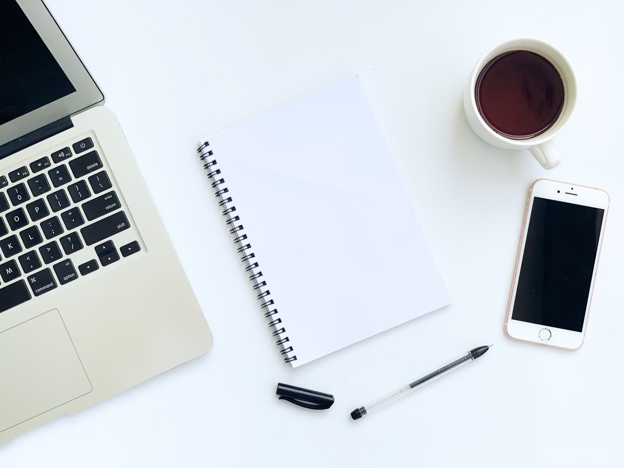 Flat lay of a clean workspace with a laptop, smartphone, coffee, and notebook on a white background.