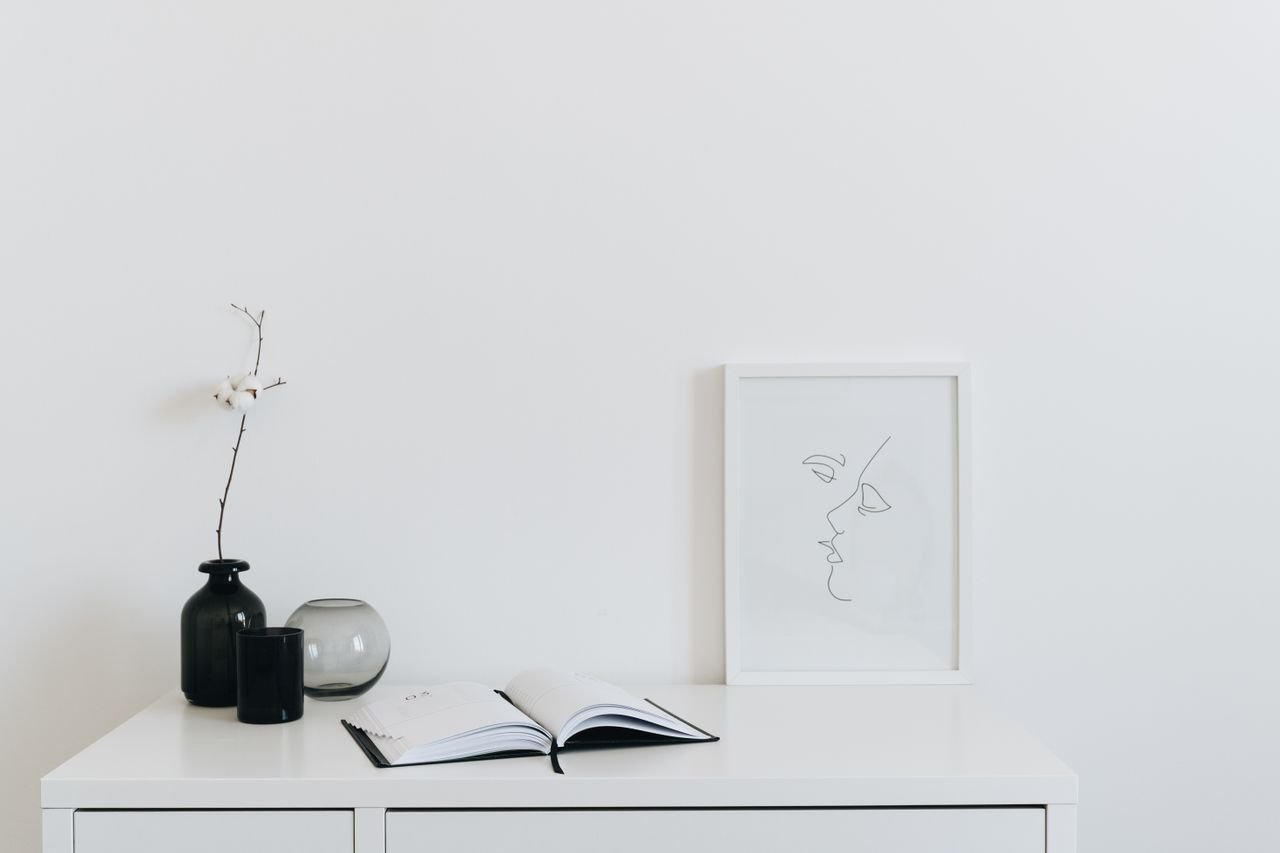 A minimalist setup featuring a desk with black vase, open book, and line art frame against a white background.
